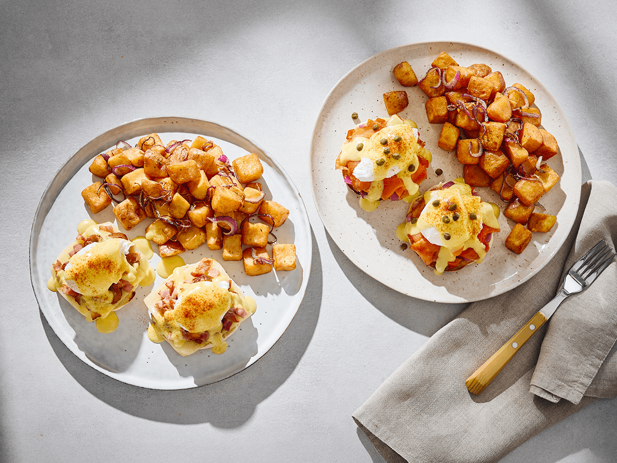 Two plates of eggs Benedict — classic and smoked salmon — with hollandaise, capers and home fries